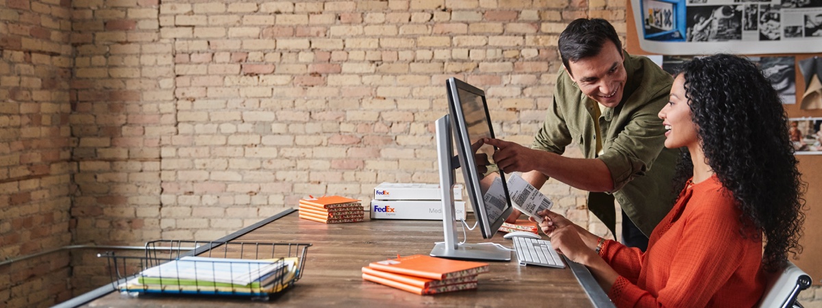 man pointing at computer monitor and smiling at a woman holding a FedEx label