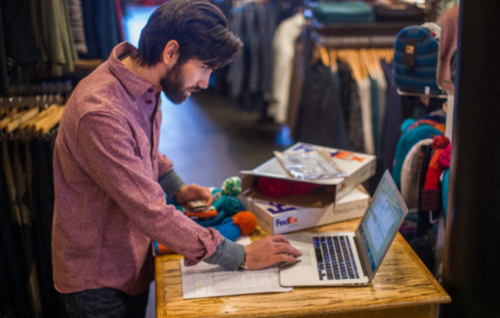 Man standing at desk looking at a laptop with an opened FedEx box next to him