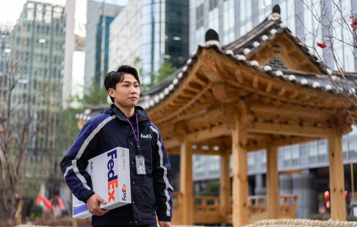 FedEx courier holding a package near a traditional wooden pavilion in an urban area with modern buildings.