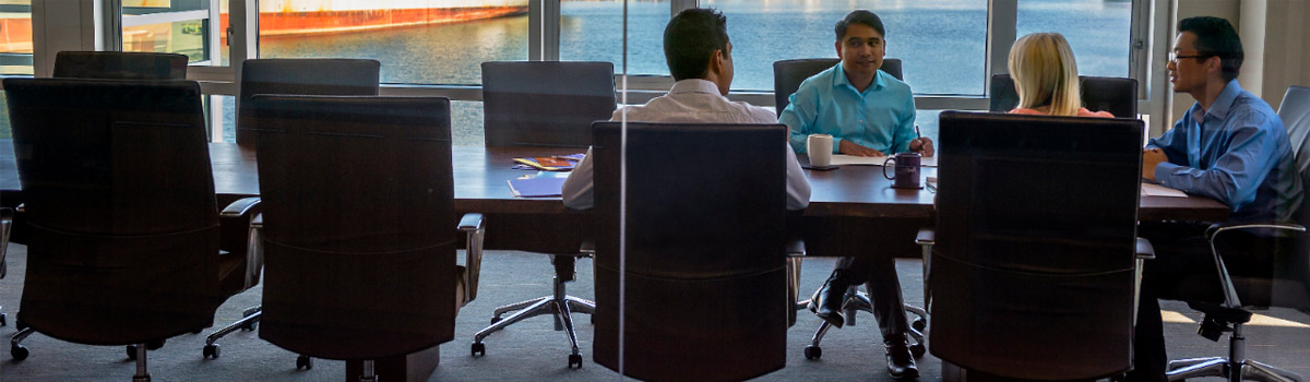 4 individuals meeting at a table with paperwork and coffee mugs, the window behind the table presents a view to water.