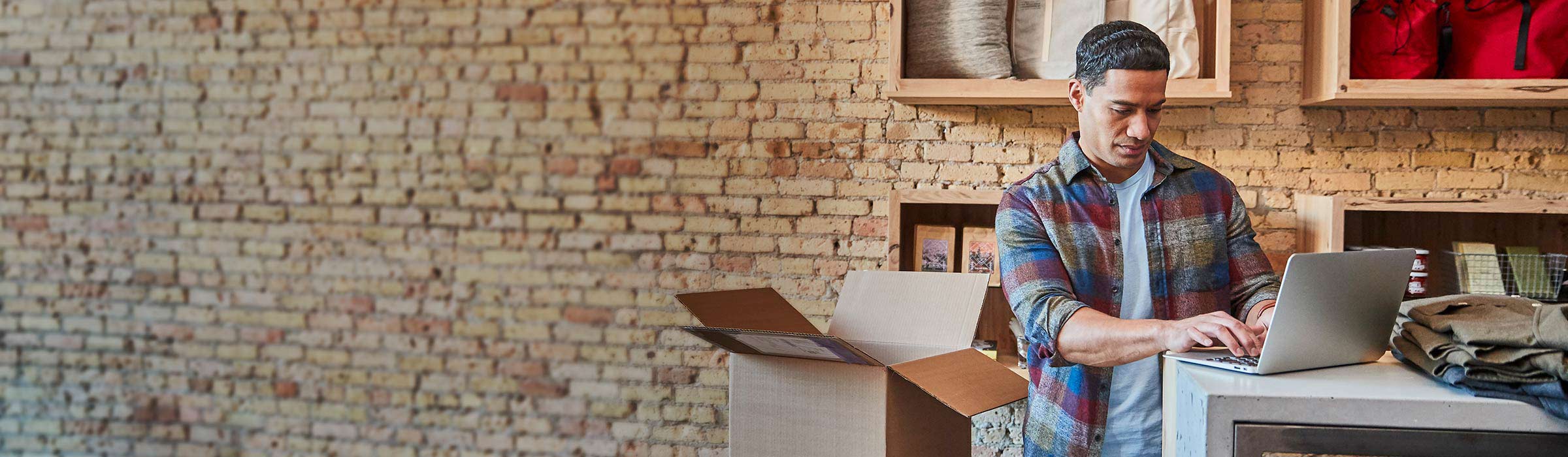 A retail shop owner works on his laptop as he unpacks inventory.