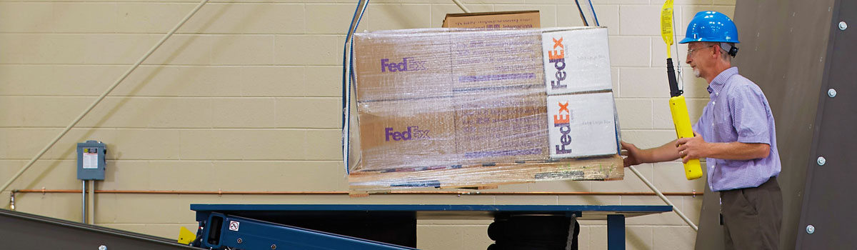 FedEx Packaging Lab employee wearing a hard hat performing a test on multiple boxes wrapped on a pallet at a table.