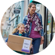 A young girl carries a box to a FedEx drop off location while her smiling mom walks behind her